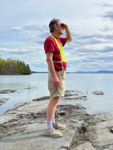 Man in red shirt and neon biking vest, shades his eyes looking from the Vermont shore across Lake Champlain.