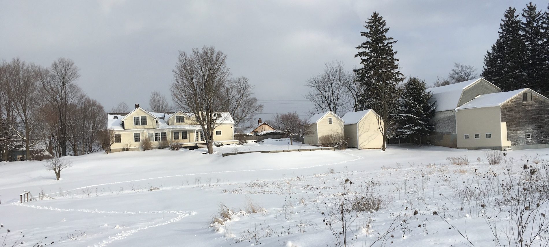 View of the yellow house and outbuildings, and a gray barn in the snow in the distance.