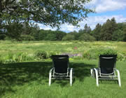 Backside of two black and white chairs in sun-dappled grass overlooking a green field with trees in the distance