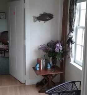 Corner of room with light wood flooring, white walls, and a wood table with lilacs in front of a window