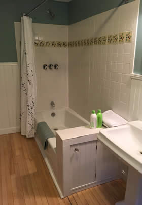 Bathroom with white tiled tub shower, white beadboard and blueish green walls, wood floors and white pedestal sink
